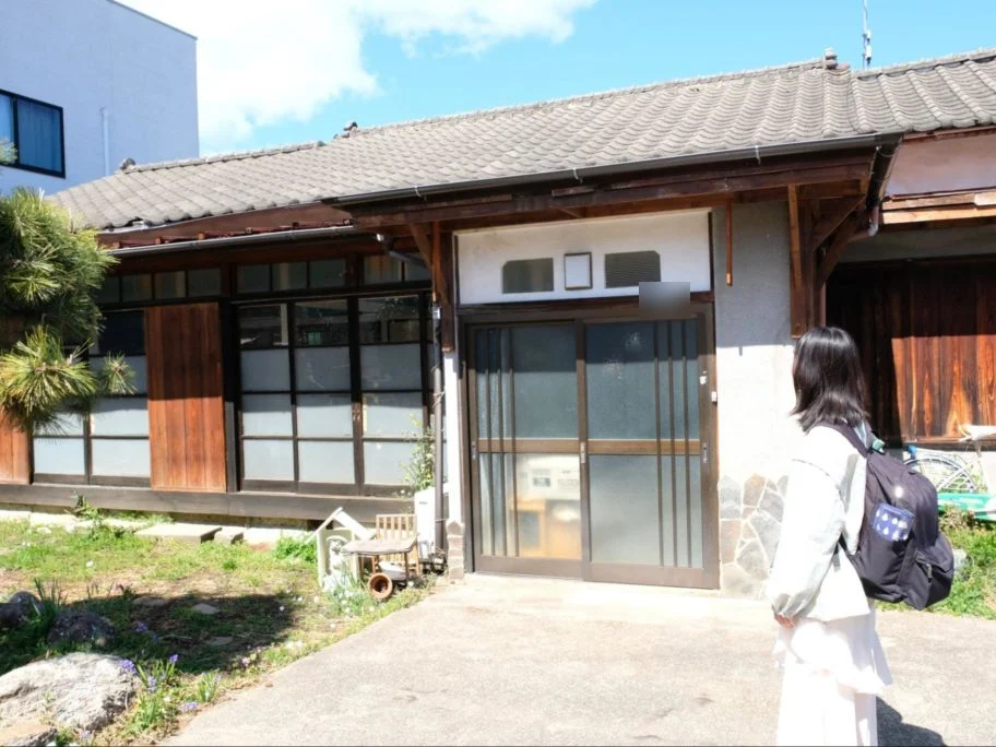 瓦屋根の平屋の前に立つ、リュックを背負った学生の後ろ姿