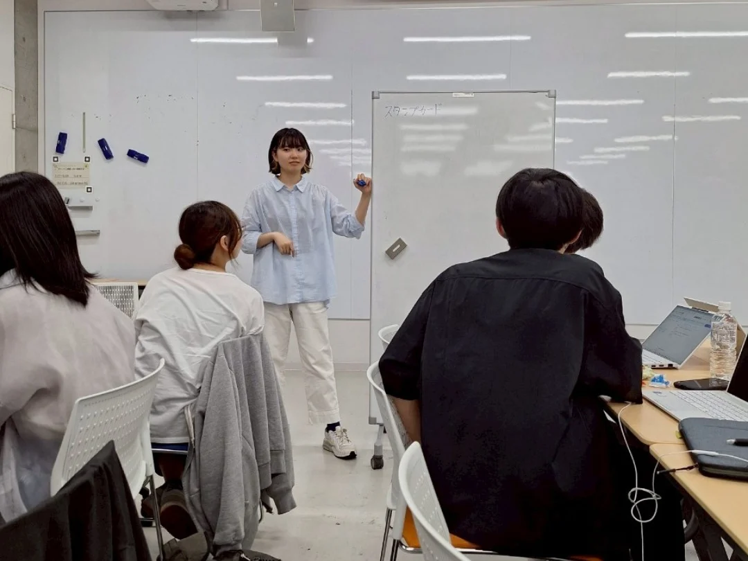 ホワイトボードの前に立つ学生と、話を聞くメンバーたち