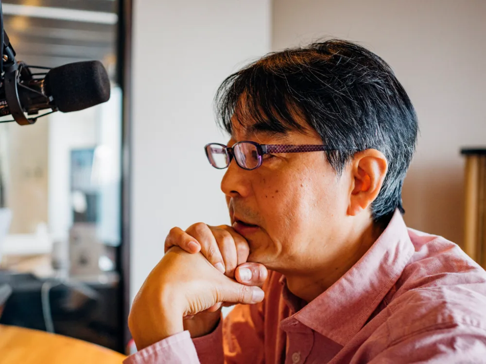 収録用マイクの前で、手を組んで考える理事長の上岡裕さん