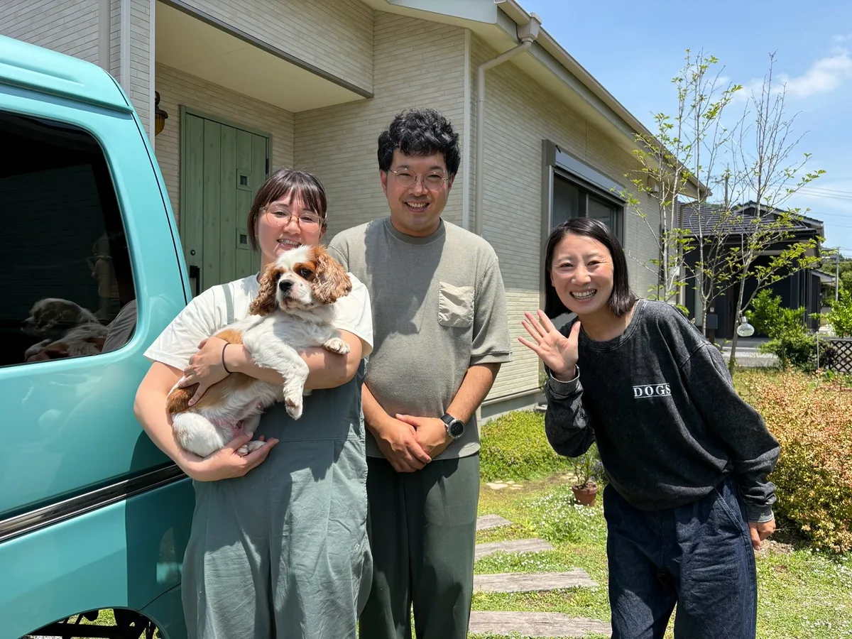 家の前で、犬を抱いた人と並んで笑顔で手をふる
