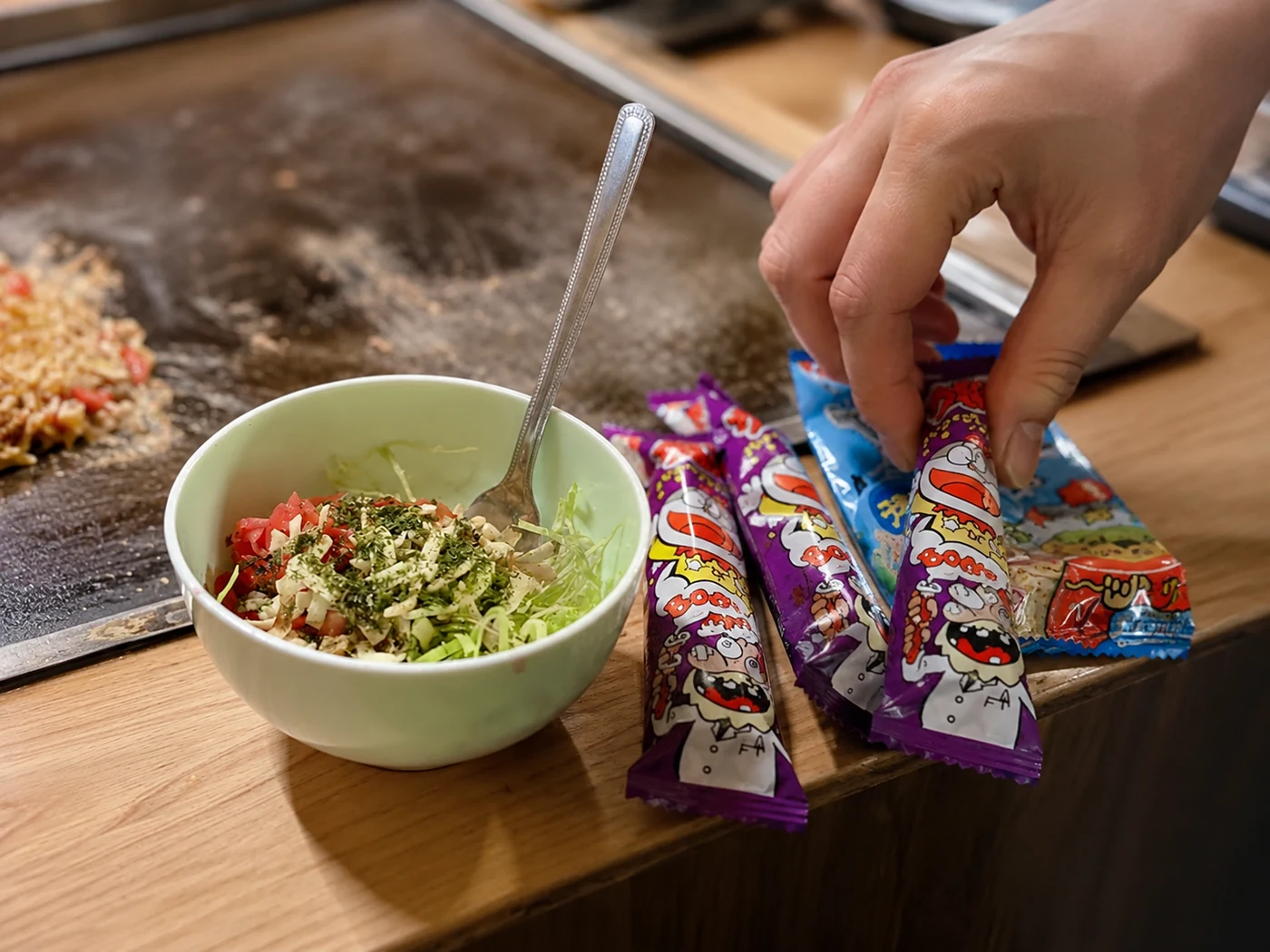鉄板の前に用意された具材の小鉢と、駄菓子を選ぶ手元