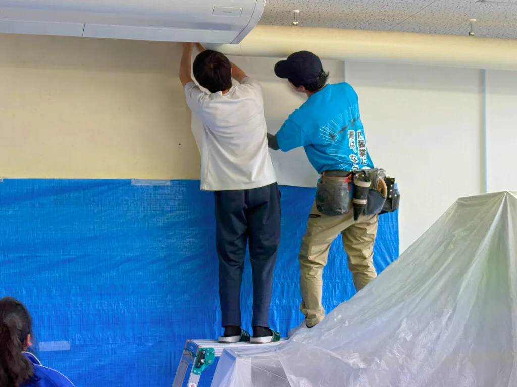 馬頭高校の進路資料室で、生徒と職人が並んで天井際のクロスを貼っている職業体験の様子