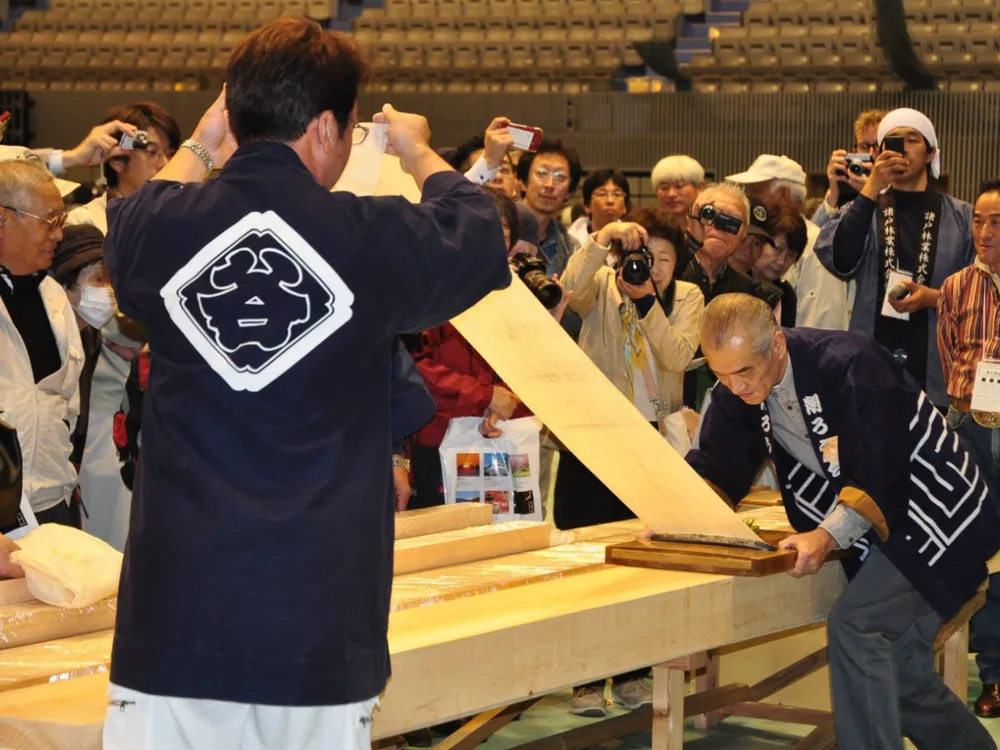 長い材から削り出した削り華を、両手で高く掲げて確かめる人と、それを見守る来場者