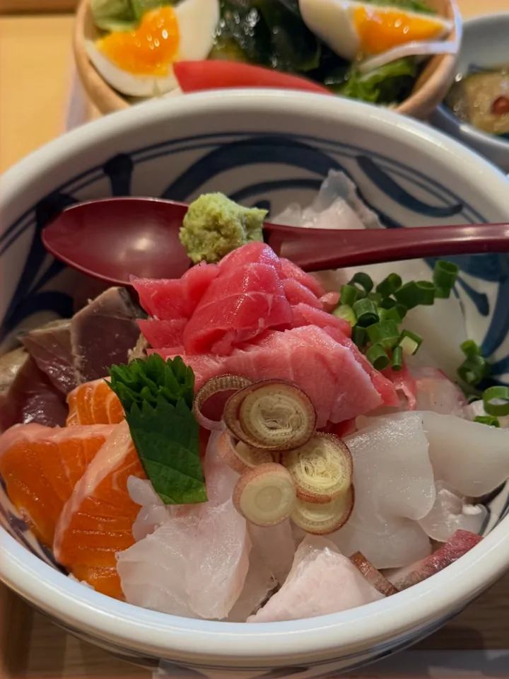 木の器に、まぐろやサーモン、白身魚、カツオがのった海鮮丼。後ろには半熟卵のサラダ