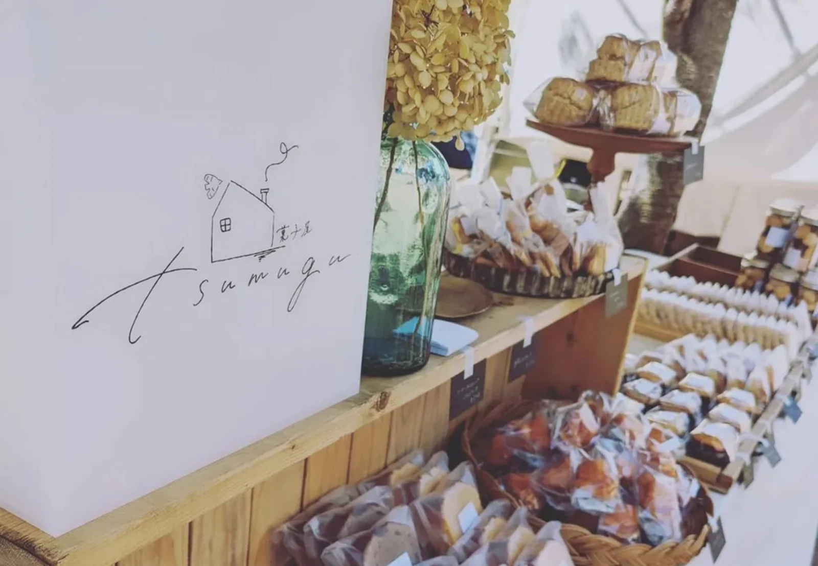 tsumugu.の文字を描いた白い看板の横に、袋詰めの焼き菓子が並ぶ棚