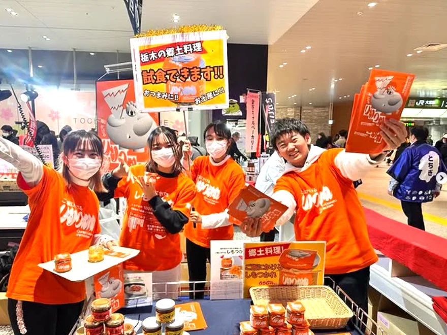 JR宇都宮駅での販売PRで、瓶詰め商品を手に笑顔を見せる栃木すみつかれ部の学生メンバー